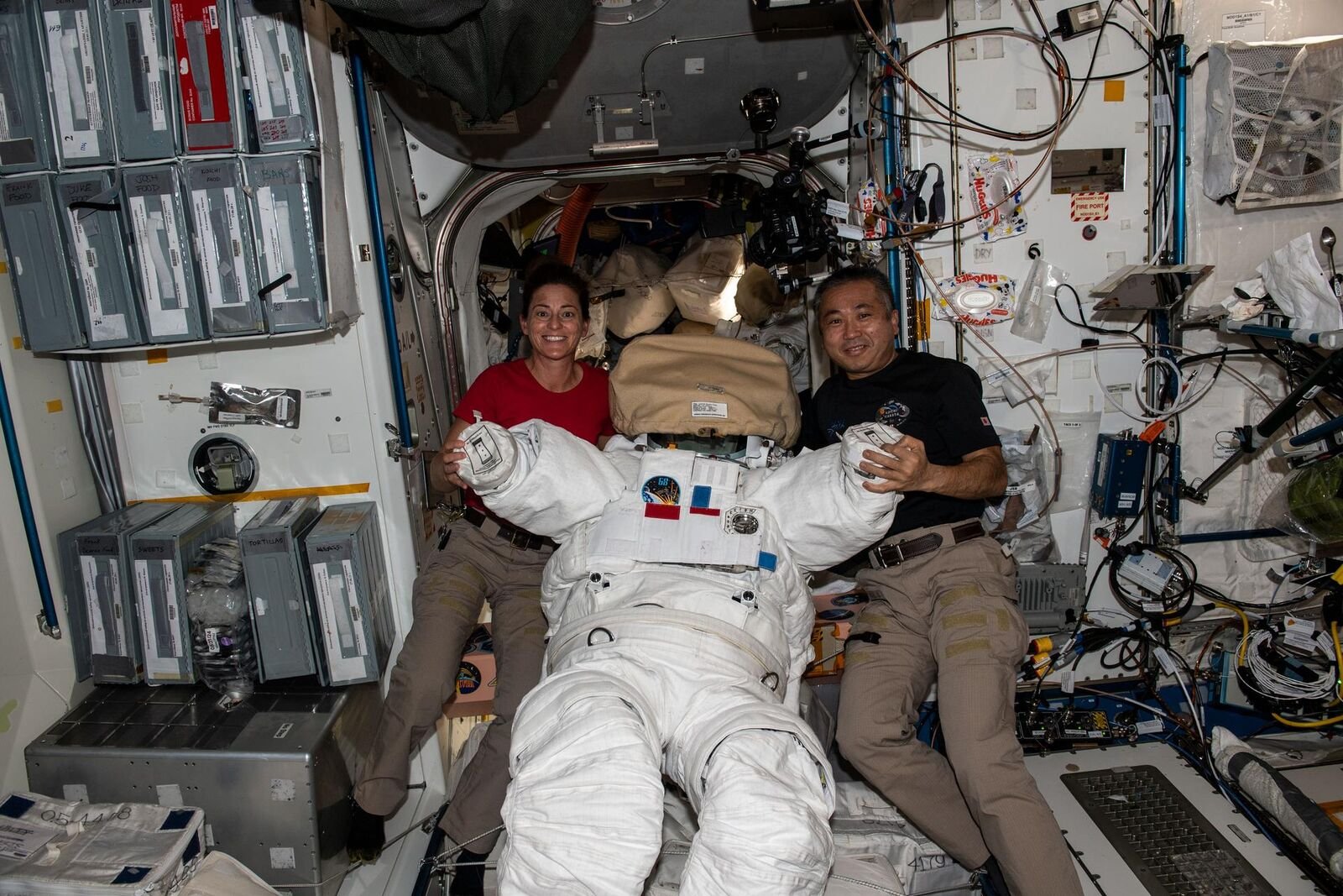 Dois trajes espaciais brancos dentro da estação espacial, diante de uma janela